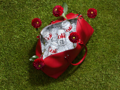 A vibrant red bag rests on lush green grass, filled with ice-chilled cans. Surrounding the bag are striking red flowers, adding a touch of elegance and contrast. The overall impression is a refreshing and summery scene, evoking relaxation and enjoyment. The composition highlights the contrast between the vivid colors of nature and the sleek design of the items.