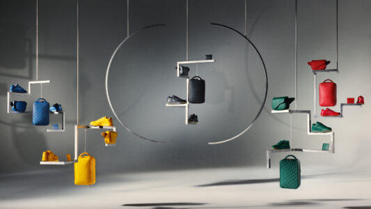 A modern display features an array of colorful shoes and bags in blue, yellow, green, and red. Positioned on minimalist, zigzagging shelves suspended from above, these fashionable accessories create a dynamic interplay of colors and shapes. Shadows and lighting add depth, highlighting the sleek, contemporary aesthetic in a harmonious and visually appealing composition.