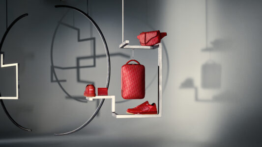 A modern, minimalist display features five red accessories: a backpack, crossbody bag, wallet, sneakers, and a small pouch. These items are carefully arranged on white geometric shelves against a soft, muted gray background. Shadows interplay with curved and straight lines, creating a sophisticated and stylish presentation. The overall composition emphasizes luxury and fashion-forward design.
