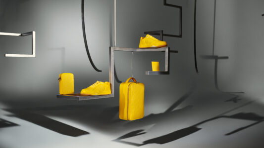 A modern grey room features various yellow accessories displayed on black suspended shelves. Items include a suitcase, a pair of shoes, a small handbag, and a cup. The striking contrast between the bright yellow and the muted background creates a stylish, minimalist aesthetic. The arrangement emphasizes the vibrant color and sleek design of each item, lending a contemporary vibe.