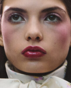 An extreme close-up focuses on a woman’s face, emphasizing glossy, deep pink lips and softly flushed cheeks. Her skin texture is natural and luminous, with visible pores and subtle imperfections. Eyes are wide and reflective, framed by minimal makeup. A delicate, patterned fabric wraps around her neck, adding softness to the intimate, tactile beauty portrait.