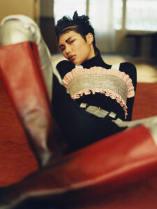 A person with short, spiky hair and a confident expression is seated, wearing a striking outfit. The attire includes a textured top over a black outfit, paired with bold, knee-high red and silver boots. The background features a warmly-lit space with soft carpet and blurred windows, creating an atmosphere of avant-garde fashion and casual elegance.