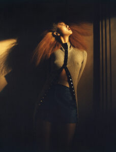 A person stands dramatically, head tilted back, with long, flowing hair lit by warm, directional light. They wear a ribbed, long-sleeved top with circular cutouts, paired with high-waisted shorts. The background features deep shadows and subtle architectural details, creating an atmosphere of intensity and style. The overall scene conveys a sense of movement, energy, and fashion-forward elegance.