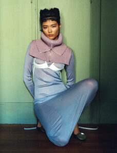 A person kneels on the floor wearing a long, light blue dress with a fitted silhouette. Over this, a structured, knit neckpiece in purple wraps around the shoulders. The outfit is accentuated by a white, futuristic bralette. The individual has short, styled hair. A mint green background and dark floor complete the unique, fashion-forward look.