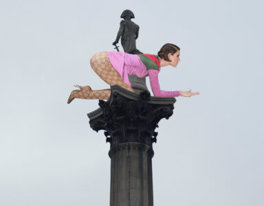 A column rises into a cloudy sky, topped with a unique statue combination. A traditional figure stands poised, holding a sword. To the side, a modern, brightly-dressed figure appears to crouch, wearing patterned boots and colorful clothing, extending a hand forward. The juxtaposition creates an unusual and striking visual contrast against the muted sky.