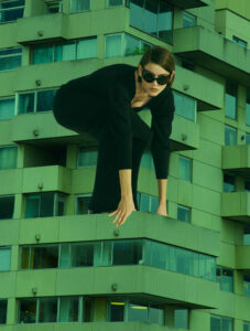 A person crouches in a striking pose, wearing dark clothing and sunglasses, giving an impression of power and agility. The backdrop is a modern building with a geometric façade composed of balconies and windows in various shades of green. The unique angle and composition create a surreal, dynamic atmosphere, blending fashion and architecture seamlessly.