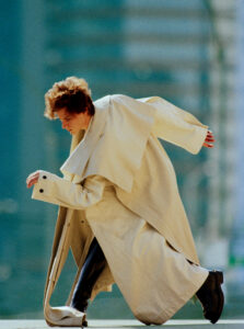 A person kneels energetically in a dynamic pose, clad in an oversized beige coat with large lapels and black boots. Their hair is tousled, and they are set against a blurred, urban backdrop with a teal hue, capturing a sense of movement and style. Shadows and light interplay elegantly, enhancing the sense of motion and contemporary fashion.