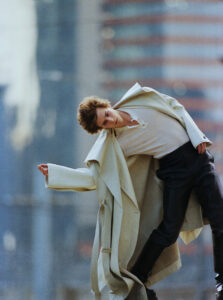 A person is captured mid-motion with one arm extended. They wear a long, cream-colored coat over a white shirt and black pants. The background features a blurred cityscape, emphasizing the dramatic pose and fashion-forward style. The overall atmosphere reflects a blend of urban sophistication and expressive movement, enhancing the modern, artistic vibe.