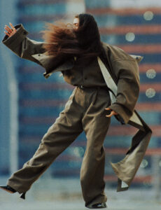 A person in an oversized brown coat and wide-legged pants strikes a dynamic pose, their long hair blowing dramatically across their face. The background features a blurred urban setting, with tall buildings and light reflections lending a sense of movement and energy. The overall atmosphere suggests a blend of fashion and urban life in motion.