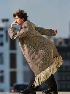 A person balances on one foot with a thoughtful expression. They wear a long, patterned coat with fringe along the hem and black footwear. The background features urban architecture, with blurred buildings and a clear sky, giving a sense of depth. The lighting casts a warm, natural glow, highlighting facial features and the coat’s intricate design.