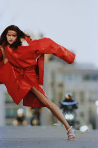 A woman stands on a street, wearing a vibrant red coat. Her pose is dynamic, leaning slightly forward, while a motorcycle with bright lights approaches from behind. Her hair flows, suggesting movement. The background features an urban setting, slightly blurred, highlighting the contrast between her vivid outfit and the neutral tones around her.