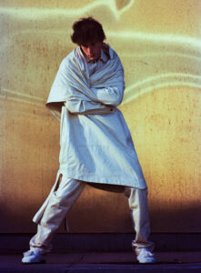 A person stands in front of a textured golden backdrop, wearing a layered white outfit. Their stance is relaxed, with arms crossed and head slightly tilted down. The ensemble includes a long, flowing coat and textured shoes. The backdrop features subtle lighting variations, adding depth and visual interest to the composition, creating an overall artistic and moody atmosphere.