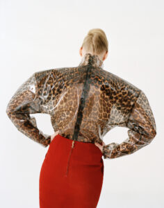 A person stands with their back turned, showcasing an outfit. The top is transparent with a leopard print pattern, accentuating the shoulders. Below, a fitted, high-waisted red skirt complements the top, with an elegant zipper detail at the back. The person's hair is styled neatly, drawing attention to the structured fashion statement against a plain background.