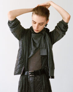 A person with slicked-back hair ties their hands behind their head. They wear a layered, dark outfit with a quilted vest, a belt, and a rolled-up long sleeve top. A scarf drapes around the neck, complementing the overall attire. Their eyes are cast downward, conveying a contemplative or serene mood against a plain background.