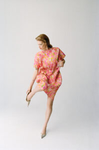 A person stands on one leg, slightly bent, wearing an elegant, colorful floral dress. The other leg is lifted with a pointed shoe partially removed. The right arm holds a transparent glass, while the gaze is directed downward. The background is minimal and pale, emphasizing the graceful and poised stance of the individual.