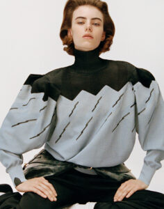A person confidently poses wearing a high-necked, patterned sweater with puffed sleeves. The garment features a zigzag design in black and light blue, creating a striking visual contrast. Their expression conveys poise and elegance. The style is complemented by dark pants and a belt, while a neutral background emphasizes the fashion-forward outfit and sophisticated demeanor.