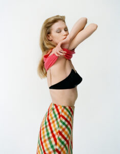 A person with long blonde hair is stretching their arms, wearing a black top and patterned skirt with red, green, and orange diagonal stripes. A pink garment is held against their chest. The background is plain white, highlighting the colors and forms of the outfit and the person's relaxed, graceful pose. Their eyes are gently closed.