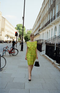 A person walks confidently down a city street, wearing a vibrant yellow dress with a subtle floral pattern. They hold a black handbag and wear pointed heels. The background showcases an elegant row of Georgian-style buildings under a clear blue sky. A red bicycle and streetlamps add to the urban charm of the scene.