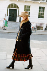 A person stands in the foreground, dressed in a stylish, dark coat with fur trim and a plaid skirt, topped with a wide-brimmed hat. They wear black heeled boots. In the background, a building with large windows has two people near the steps, and a red bicycle is parked beside a railing on the sidewalk.