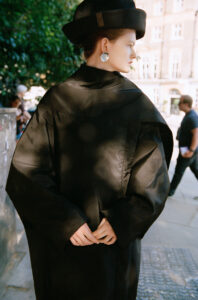 A person stands in profile, adorned in elegant black attire and a matching wide-brimmed hat. Silver earrings reflect the daylight, emphasizing their sophisticated look. Hands are gently clasped at the back. The scene is set outdoors, where soft dappled sunlight filters through leaves, casting gentle shadows. Blurred figures and a building complete the urban backdrop.