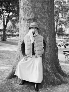 A person leans against a large tree in a park, dressed in a fuzzy, buttoned vest over a long skirt and boots. Their head is adorned with a wide-brimmed hat and a scarf, adding an air of vintage elegance. The background is softly focused, revealing a serene park setting with scattered benches and dappled sunlight filtering through the leaves.