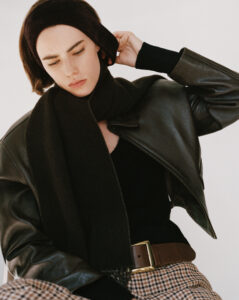 A person with short hair wears a dark headband and a matching scarf, paired with a leather jacket. The individual seems to be adjusting the headband while displaying a focused expression. Their outfit includes a brown plaid skirt, a black top, and a dark belt with a golden buckle. The background is minimalist, enhancing the stylish ensemble.