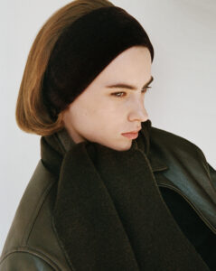 A person faces right, wearing a dark headband and an olive green coat. Their hair is parted, and they're wrapped in a thick, dark scarf. The soft lighting highlights their contemplative expression and the texture of the clothing. The background is a simple, light tone, putting emphasis on the subject's features and attire.