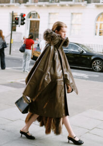A person wearing a long brown coat with a large fur-trimmed hood walks on a city sidewalk. They carry a black handbag and wear black heels. The background features a street scene with a pedestrian crossing, a black car, and buildings with arched windows. A small group of people is visible, including one wearing a red shirt.