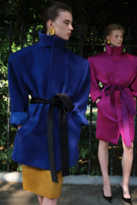 Two individuals stand confidently outdoors. One wears a vibrant blue coat with a high collar, accented with a black belt and gold earrings, paired with a mustard skirt. The other is dressed in a striking magenta coat, similarly belted, with black heels. A blurred, lush background of foliage and iron railings adds depth and contrast to the scene.