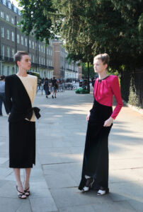 Two individuals stand on a sunlit city sidewalk, surrounded by classic architecture and greenery. They wear elegant outfits, one in black and white, the other in black and pink, both with long skirts and stylish shoes. Bicycles and pedestrians are visible in the background, and large trees cast dappled shadows on the pavement, creating a serene urban scene.