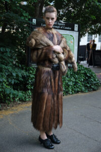 A person stands confidently on a paved path surrounded by lush greenery. They're wearing a long, luxurious fur coat with a subtle blend of brown tones. Black shoes add elegance and a small black bag is slung over the coat. The background features people in conversation, a detailed map, and the presence of leafy trees.