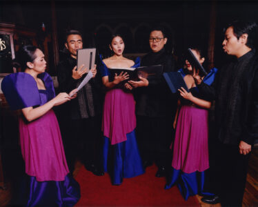 A group of six individuals stands in a semicircle, appearing to sing. Three women wear purple and pink dresses, while the men are dressed in black formal attire. Each person holds a music book or sheet. They are situated in a richly decorated room, suggesting a formal or performance setting. Warm lighting highlights their expressions and gestures.