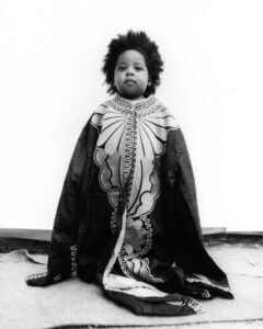 A young child sits with a calm expression, enveloped in an oversized, intricately patterned shawl. The monochrome setting highlights the detailed floral and geometric designs of the cloth. Their hair is styled naturally, adding to the serene and contemplative mood. The backdrop is minimalist, ensuring focus remains on the child and the elaborate textile wrapped around them.
