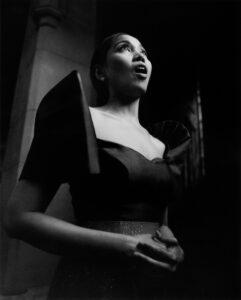 A woman stands dramatically against a dimly lit background, exuding elegance and focus. She wears a dress with striking, angular sleeves, gazing upward with an expression of passion. Her hands are clasped at her waist, emphasizing her poised and graceful demeanor. The soft shadows and highlights create a mood of intensity and anticipation, evoking a moment of profound emotion.