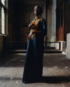 A person stands in an ornate setting, wearing a long, dark robe with intricate golden patterns. They hold a bright yellow flower, with a relaxed posture. The lighting creates dramatic shadows, highlighting their glasses and layered necklaces. The background features large windows and detailed stonework, suggesting a historical or ecclesiastical environment, enhanced by the ambient natural light.