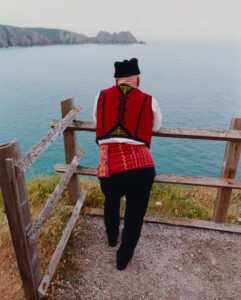 A person stands on a coastal path, overlooking a vast, serene body of water. They wear a traditional costume with a vibrant red vest and intricate yellow and black patterns. The headgear is black, and the surroundings include wooden fencing and grassy terrain. In the distance, rugged cliffs meet the calm sea under a soft, overcast sky.
