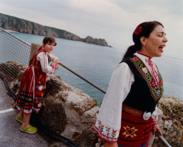 Two individuals stand near a seaside, dressed in vibrant, traditional attire with intricate embroidery. The background features a vast, calm ocean with distant cliffs under a cloudy sky. One person appears to be singing or speaking passionately, while the other looks relaxed, leaning on a stone wall. Their clothing includes vivid patterns and a rich color palette.