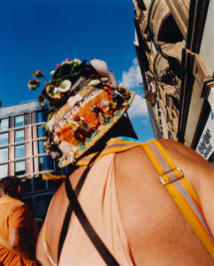 A person wears a vibrant hat adorned with small figurines and decorations. Straps cross over the person's back, with a clear sky and urban architecture in the background. The surrounding buildings display a mix of modern and traditional styles, creating an interesting contrast. People nearby add to the lively atmosphere of the scene.