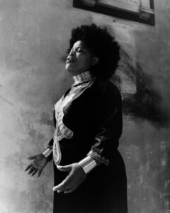 A woman stands with expressive posture against a textured background, her eyes closed as if deeply immersed in emotion. She is wearing a long, flowing dress adorned with intricate patterns and metallic embellishments. Her hair is styled in a voluminous afro. The setting appears to be indoors with a rough, aged wall adding a rustic atmosphere.