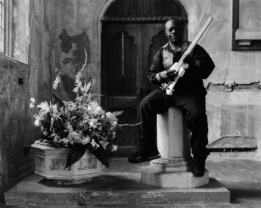 An individual sits on a pillar, holding a guitar. Their attire is dark, featuring a patterned shirt. They are situated in a rustic setting with textured walls and an arched wooden door. Beside them, a large vase holds a vibrant array of flowers, adding contrast and elegance to the otherwise muted environment.