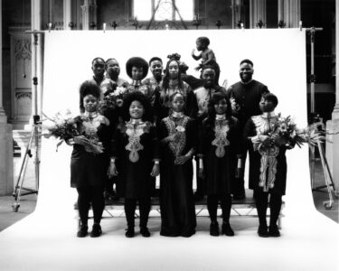 A group of fourteen individuals of varying ages poses together, dressed formally in dark clothing. Several hold bouquets of flowers, while two children are included among the adults. The backdrop appears to be a simple white screen set against a darker, detailed room with columns, adding contrast to the scene. The atmosphere feels celebratory and elegant.