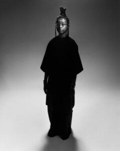 A person stands confidently, wearing a black, flowing outfit in a studio setting. Their hair is styled upward with intricate braids, adorned with beads. Large round glasses frame their face, complemented by bold makeup featuring dots above the eyebrows. Metallic bracelets accentuate the wrists, adding an artistic touch. The lighting creates a strong contrast, highlighting their unique style.