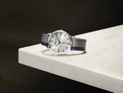 A sleek, classic wristwatch with a silver dial and minimalist design features thin, black hour markers and a pair of slender hands. The watch is secured with a dark leather strap, placed elegantly against a polished marble surface. Its refined appearance and subtle elegance suggest a timeless piece with understated sophistication, perfect for a sophisticated setting.