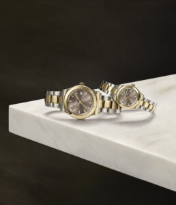 Two elegant wristwatches with metal bands are displayed on a smooth, glossy white marble surface. The watches combine gold and silver tones, featuring dark dials with sleek markers. Positioned diagonally, they reflect light subtly, adding a luxurious touch. The background is a dark, muted shade, emphasizing the timepieces' sophisticated design and craftsmanship.