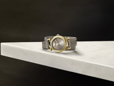A sophisticated timepiece rests on a smooth, white marble surface with subtle veins. Its round, gold-colored case and silver-hued dial are complemented by a chic, taupe leather strap featuring delicate stitching. The background is a deep, rich black, enhancing the luxurious feel. Soft lighting accentuates the watch's elegant design and craftsmanship.