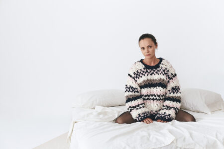 A person with a calm expression sits on a neatly made bed in a simple, bright room. Wearing a cozy, oversized sweater with a chunky knit pattern in soft colors, they are surrounded by white pillows and linens. The atmosphere is serene and minimalist, focusing on the contrast of textures and gentle colors.