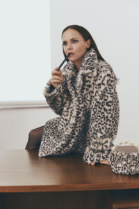 A person with long, dark hair sits on a wooden surface, holding a pen near their lips in a thoughtful pose. They wear a plush, leopard-print coat, and black stockings. A matching leopard-print accessory with a braided handle is placed beside them. The setting has a minimalist feel, with white walls and ample natural light.