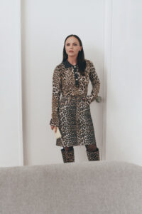A woman stands confidently near a white wall and door, wearing a leopard print dress and matching knee-high boots. Her long, dark hair is worn straight, and she carries a small beige clutch. Her expression is poised and self-assured. In the foreground, the edge of a gray sofa can be seen, adding depth to the setting.