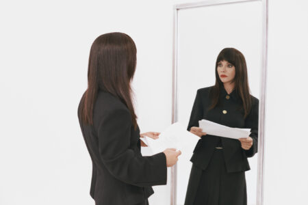 A woman, dressed in a black outfit with gold buttons, stands holding papers in front of a full-length mirror. Her dark, straight hair frames her face as she gazes at her reflection. The minimalist setting features a stark, white background and an empty room, creating a focused and introspective atmosphere surrounding the solitary figure.