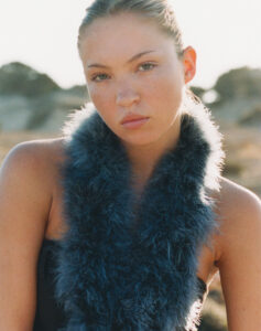 A young woman with light skin, freckles, and light brown hair pulled back wears a strapless top with a fluffy, dark blue scarf draped around her neck. Her expression is calm and neutral, and the background is softly blurred, suggesting an outdoor setting with natural light illuminating her features, casting gentle shadows on her face and shoulders.